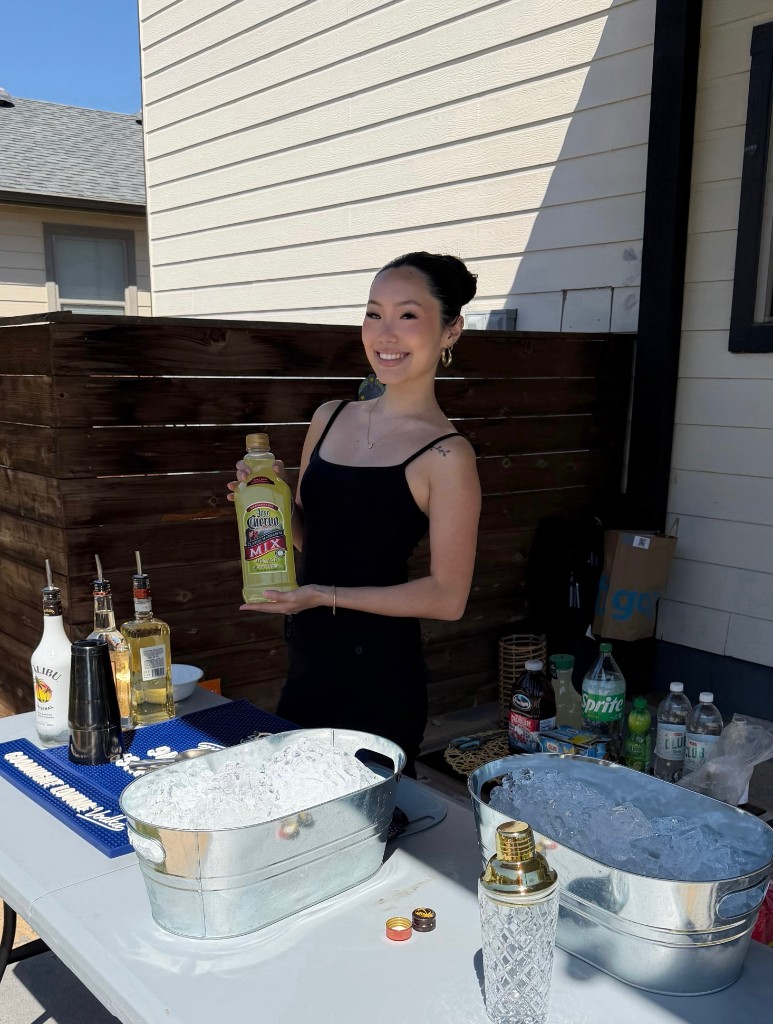 Smiling female Houston bartender holding Jose Cuervo margarita mix at a backyard party with ice buckets and bar setup
